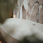 Banheira branca com espuma e torneira cromada com água corrente em banheiro aconchegante com velas e roupão branco