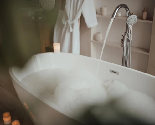 Banheira branca com espuma e torneira cromada com água corrente em banheiro aconchegante com velas e roupão branco