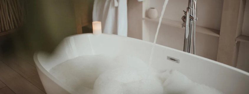 Banheira branca com espuma e torneira cromada com água corrente em banheiro aconchegante com velas e roupão branco