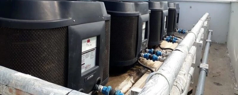 Sistema de bombas de calor instalado em um espaço externo, mostradas alinhadas ao longo de uma parede, com tubulações de água e conexão ao sistema de aquecimento ou resfriamento.