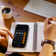 Pessoa usando calculadora no celular e anotando números em caderno espiral ao lado de teclado e xícara de café