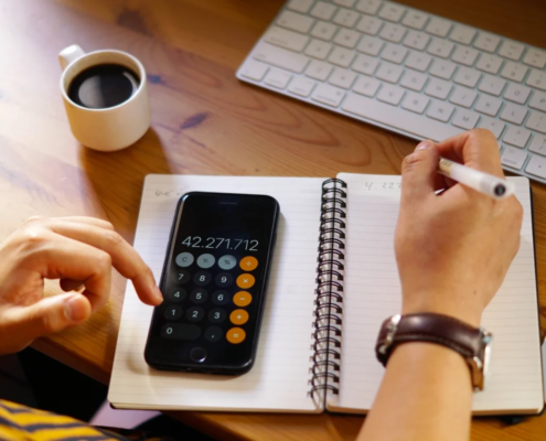 Pessoa usando calculadora no celular e anotando números em caderno espiral ao lado de teclado e xícara de café