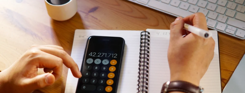 Pessoa usando calculadora no celular e anotando números em caderno espiral ao lado de teclado e xícara de café