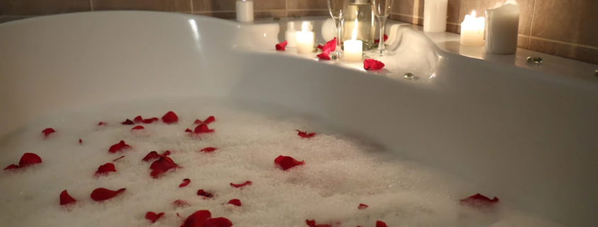Banheira de hidromassagem com bolhas de sabão, pétalas de rosa, velas acesas e taças de espumante, criando um ambiente relaxante e romântico.